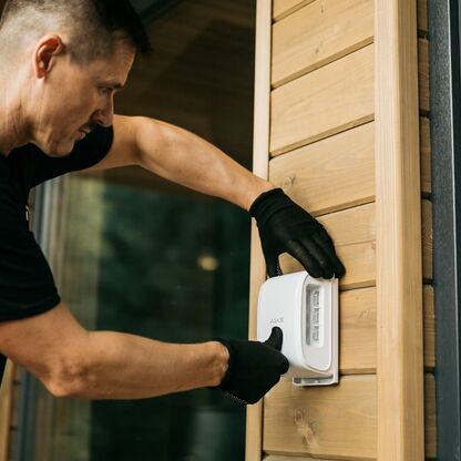 Ein Techniker installiert an einer Holz-Außenwand eine Einbruch­melde­anlage